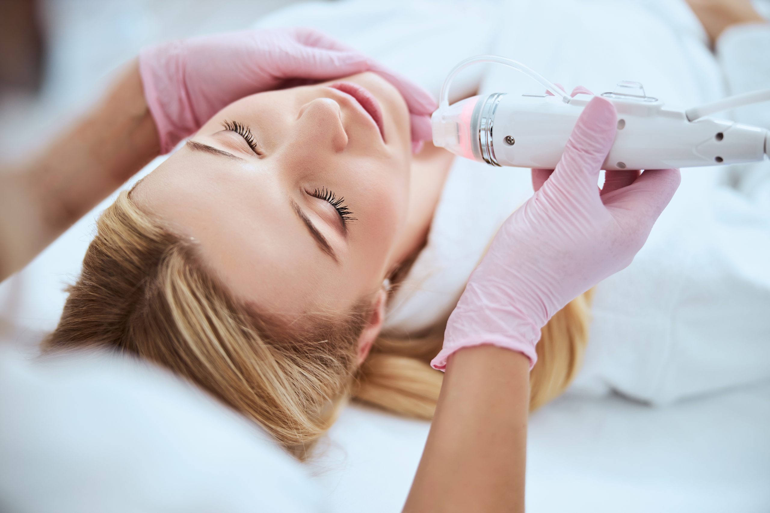 A woman lying back with her eyes closed while gloved hands guide a white handpiece with a glowing tip and a thin tube along her jaw, a photo from Bellava’s blog on facials