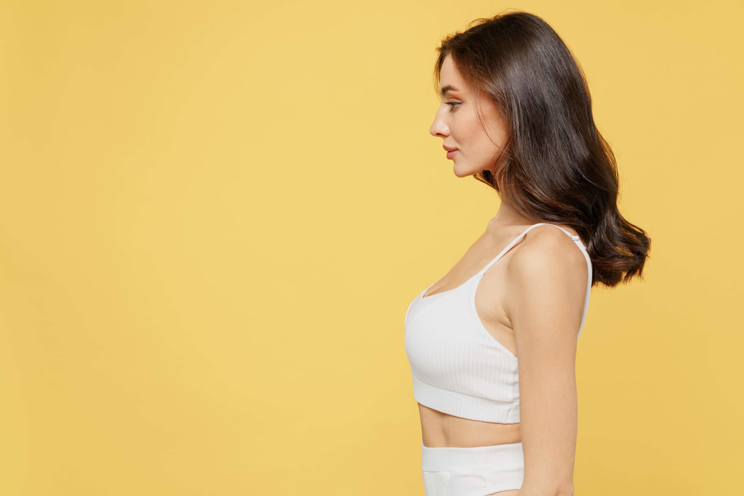 A dark-haired woman in a white ribbed crop top, seen in profile against a plain yellow background