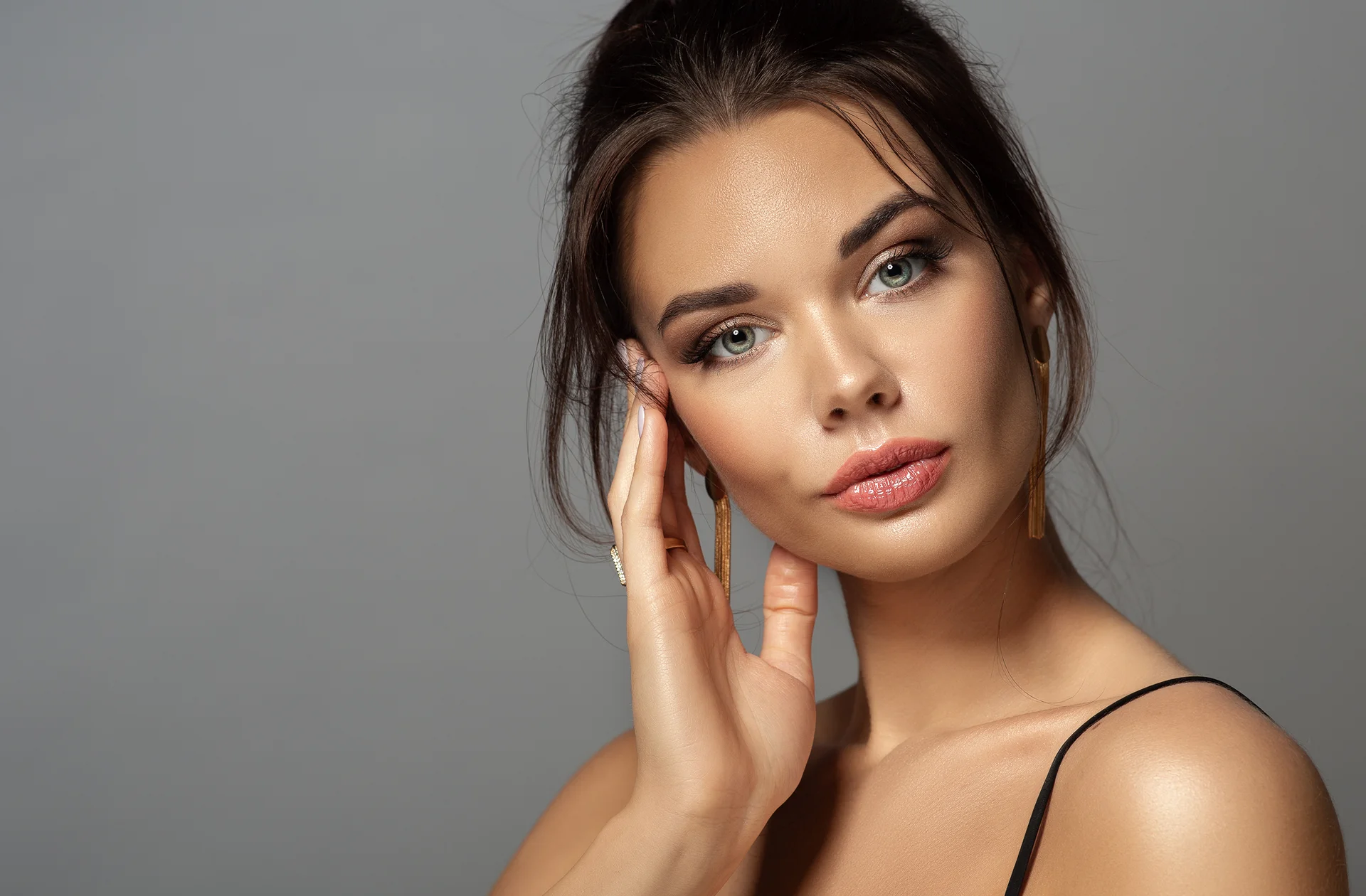 A dark-haired woman with gold drop earrings resting her fingertips at her temple, from Bellava’s blog on natural-looking lip fillers