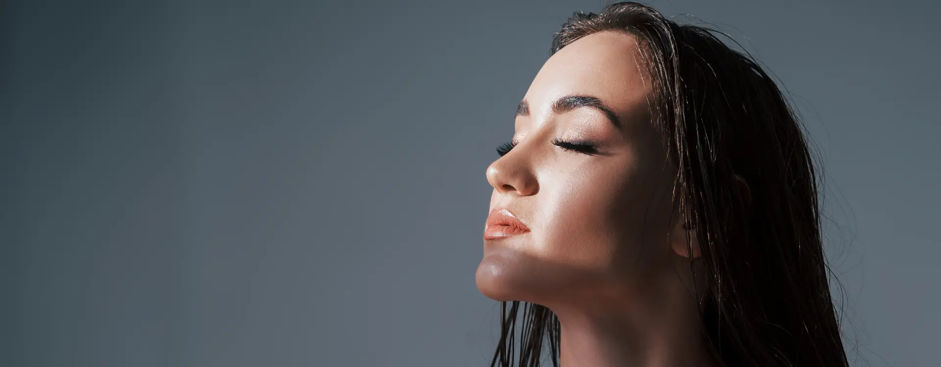 A woman with long dark hair and closed eyes tilting her face up into the light against a grey backdrop, the photo on Bellava’s photofacial page